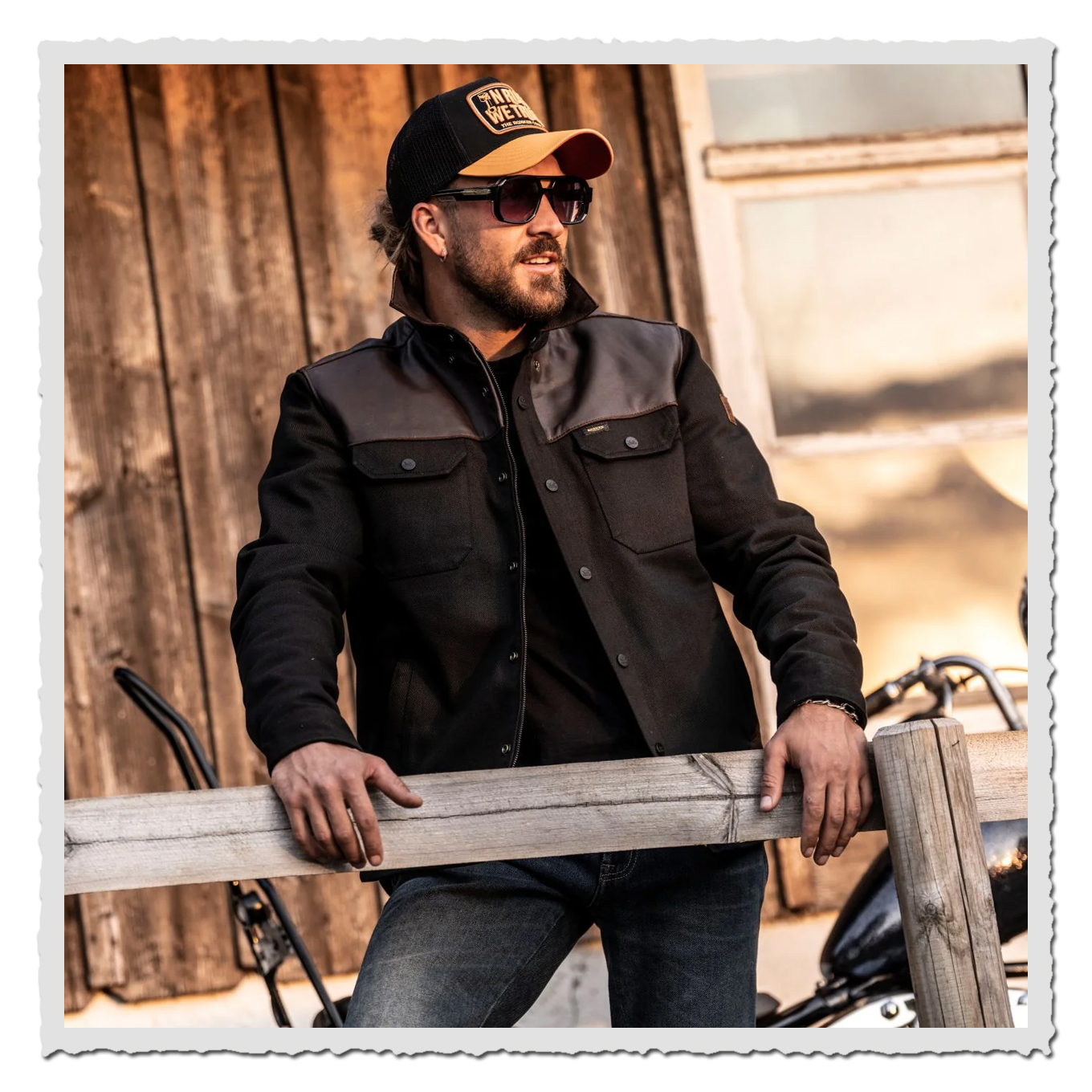 Motivfoto: Mann mit Kappe und Sonnenbrille trägt schwarzes Rider Shirt, lehnt an Holzzaun vor Holzwand.